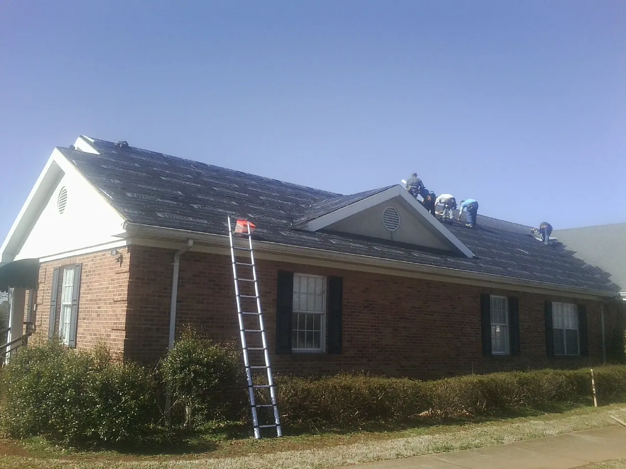 Crew tearing shingles off a brick ranch roof with a ladder set — Emergency Roof Repair work by our crew in Heath