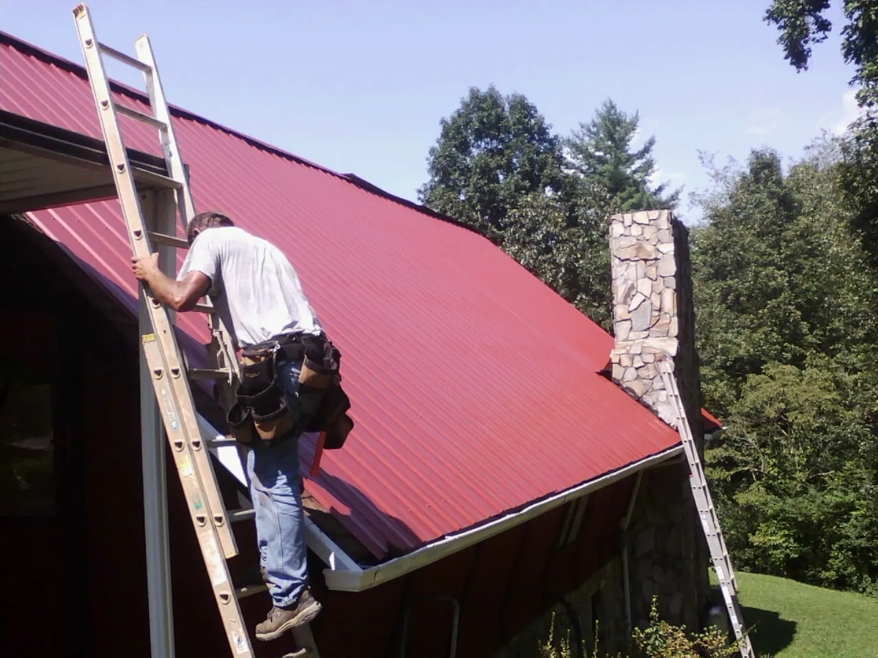 Roofer on a ladder beside a red metal roof and stone chimney — Soffit & Fascia Repair by Roofing Brothers in Heath